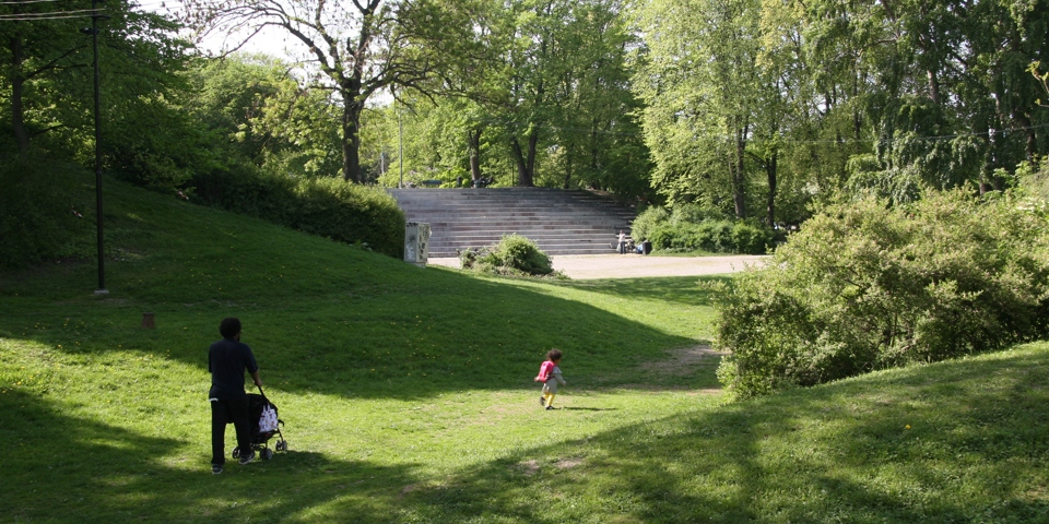 En man och ett barn går genom en grönskande park mot en utescen med läktare.