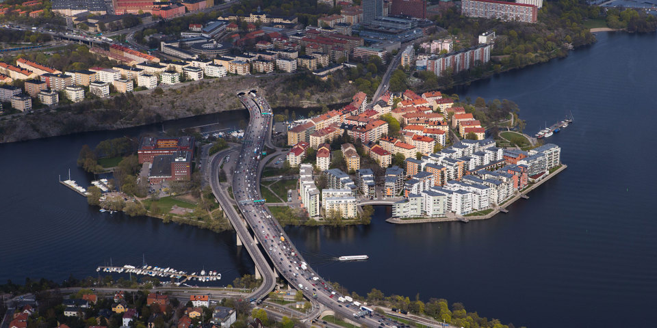 Flygvy över Lilla Essingen. Vägar med rörlig trafik. Bostadsområden och verksamhetsområden. Vatten och båtplatser vid kajer och strandkanter. Foto.