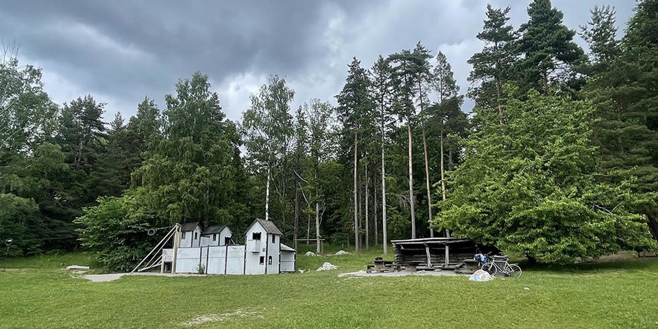 Vy mot lekplats med grillplats, lekställning i form av ett hus och linbana i ett skogsbryn, fotografi.