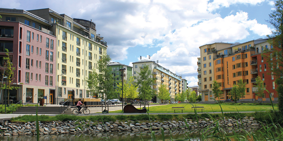 Gräsmatta bakom en vik med flerbostadshus i bakgrunden. En person som cyklar, foto.