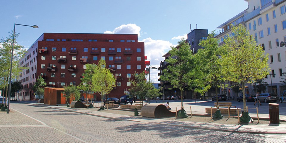 Ett stenbelagt torg med träd och sittplatser,. Omgivet av flerbostadshus, foto.
