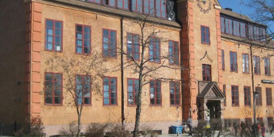 Röd skolbyggnad, skymt av skolgård och barn i rörelse, fotografi.