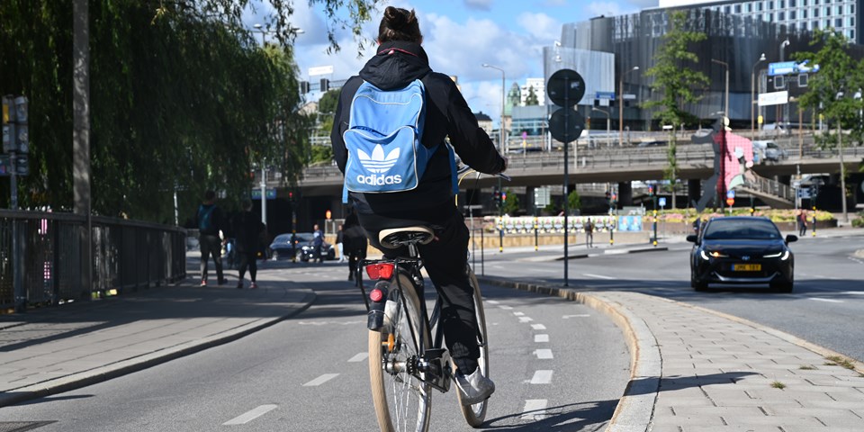 Cyklande person med ryggsäck sedd bakifrån på en cykelbana. I bakgrunden syns en bil, byggnader och människor i rörelse, fotografi.