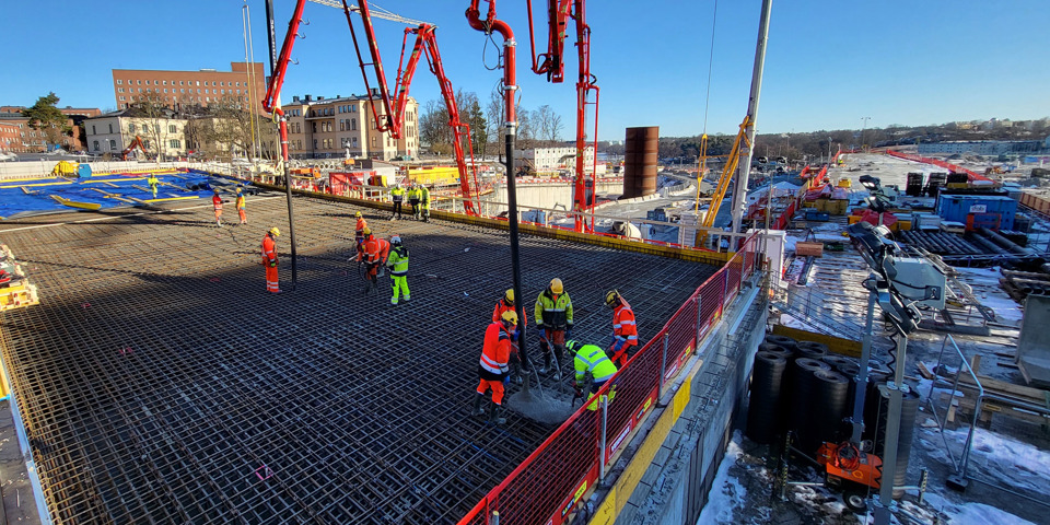 Byggarbetare i arbetskläder gjuter en betonggrund på en stor byggarbetsplats med kranar och utrustning under en klarblå himmel.