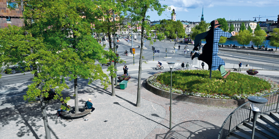 Ett stenbelagt torg med träd och en rund gräsyta i mitten. På gräsytan finns ett konstverk, foto.