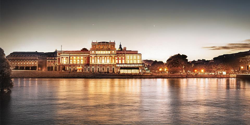 Visualisering av Kungliga Operan i Stockholm med en föreslagen påbyggnad, kvällstid sett från Slottskajen. Byggnaden är upplyst och speglas i vattnet.