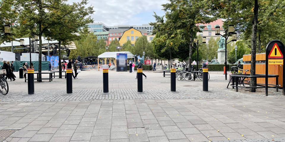 Trafikhinder framför torg. I bakgrunden träd,  byggnader, en scen, människor i rörelse, fotografi.