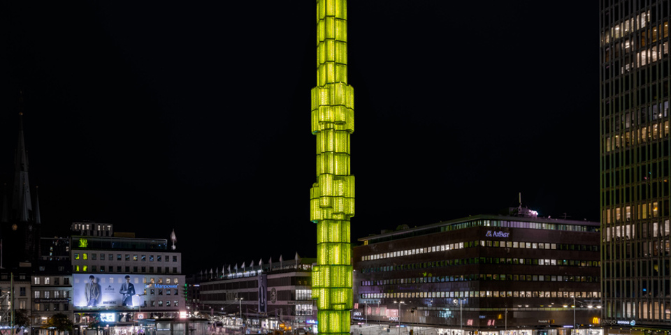 Obelisk upplyst i gult i mitten av en fontän, kväll. Byggnader och neonskyltar i bakgrunden, storstadsmiljö, fotografi.
