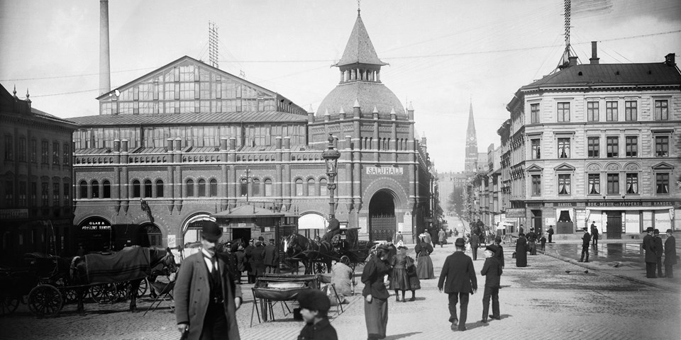 Östermalms saluhall i bakgrunden.  I förgrunden rör sig 1890-talets stockholmare., foto.
