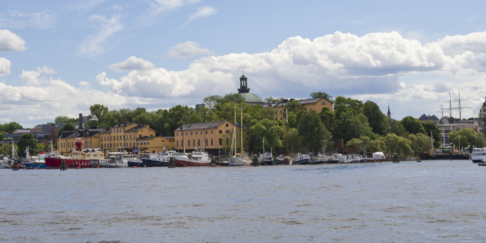 Båtar, byggnader och träd under en blå, molnig himmel. Vy från vattnet.