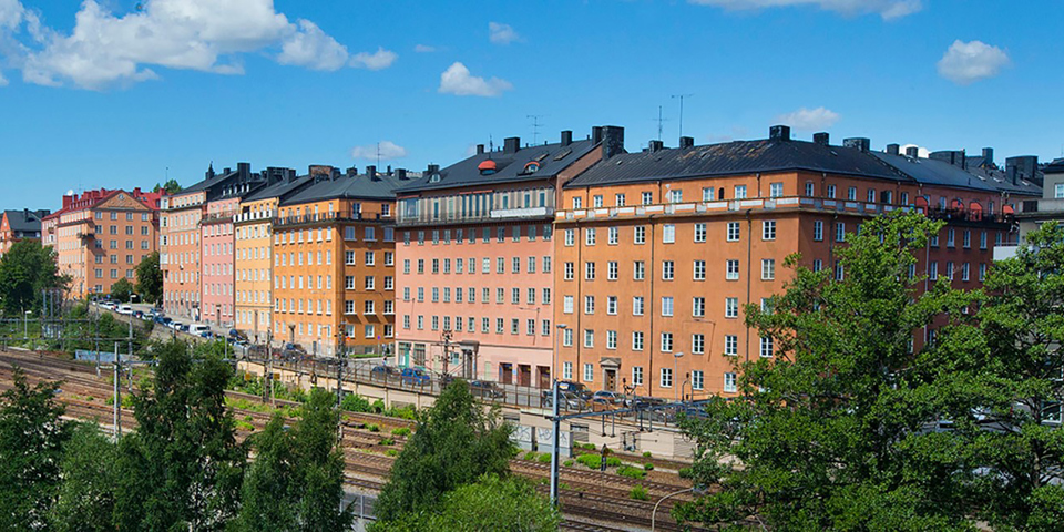 Bostadshus i flera olika färger. Omgivande träd, tågspår och gator med bilar. Foto.