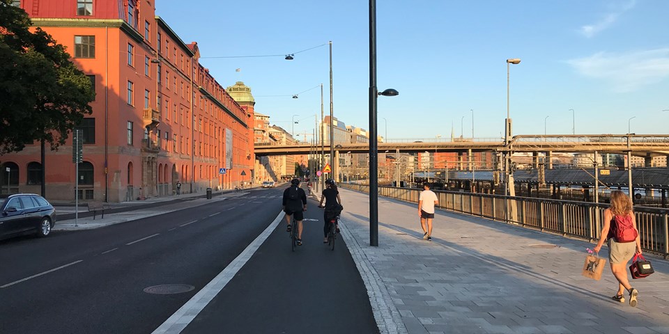 Cykelbana, bilväg och trottoar, vy mot bro i bakgrunden. Byggnad, tågspår och människor i rörelse, fotografi.