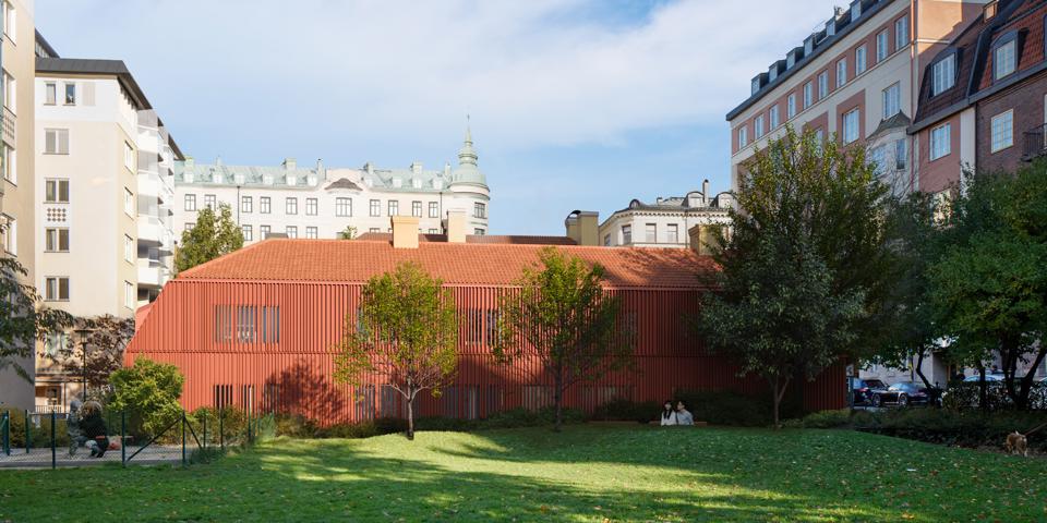 Den tänkta flygelbyggnaden i faluröd panel. Sedd från Monica Zetterlunds park med gräs och träd i förgrunden.