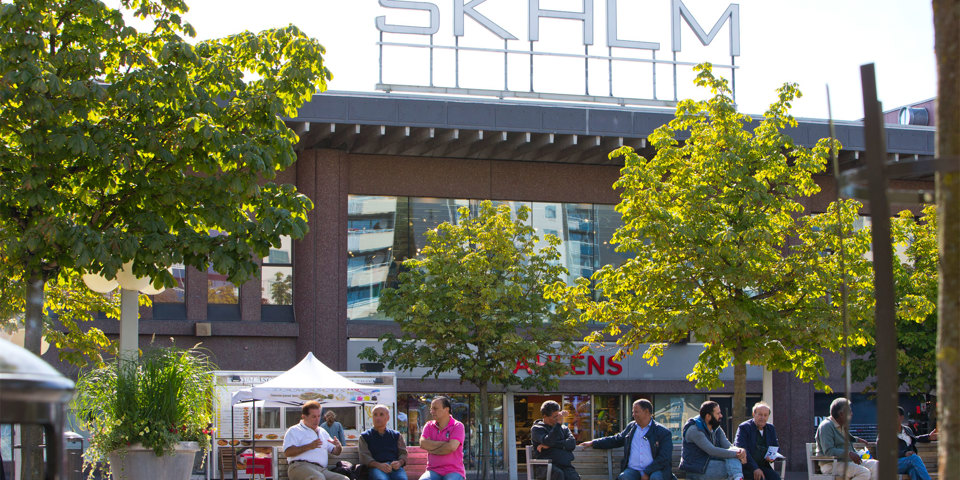 Torg med människor på bänkar. Byggnad i bakgrunden med stor skylt med versalerna "SKHLM" på taket.