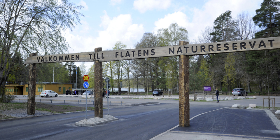 Infart till parkering vid naturreservat. Skylt i trä ovanför infarten  där det står "Välkommen till Flatens naturreservat".