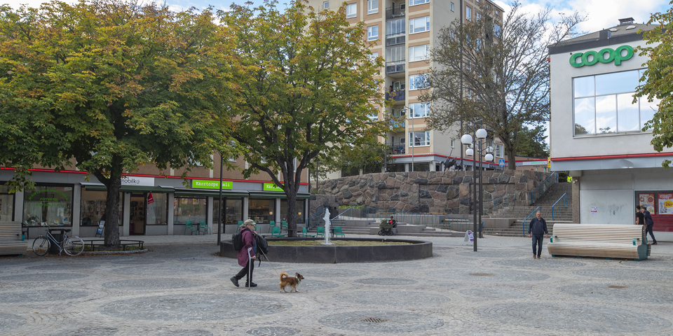 Torg, butiker, flerbostadshus, träd, människor i rörelse, fotografi.