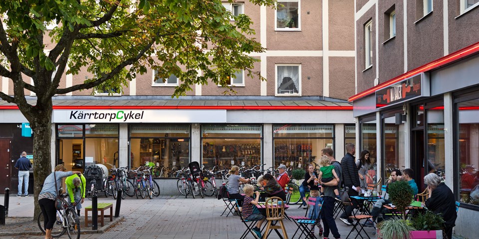Torg med butik och restaurang. Träd, bordar och stolar. Människor som äter på restaurangen. Cykelställ med cyklar och människor i rörelse. Foto. 
