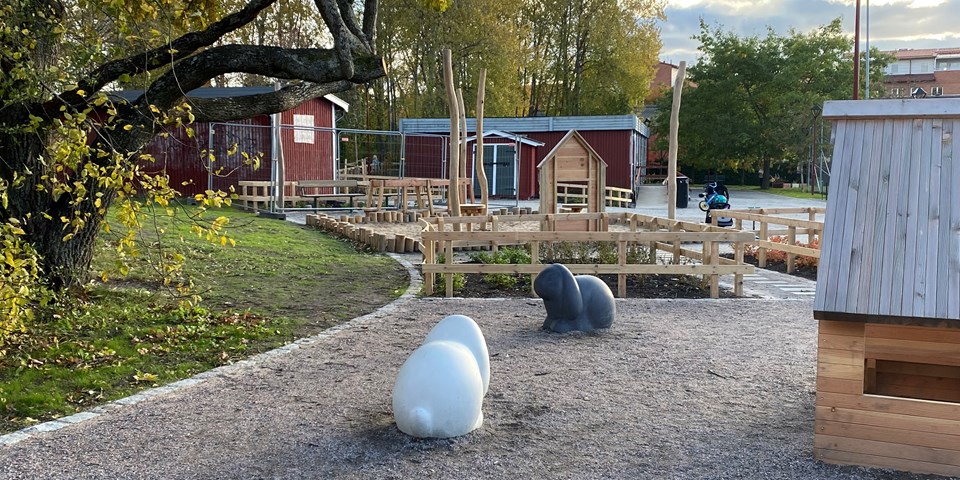 Lekplats med lekhus, kaninstatyer, gångbro i träd, staket runt planteringar, sandlåda, stora träd, sittplatser och en gångväg fram till ett litet rött hus. Foto
