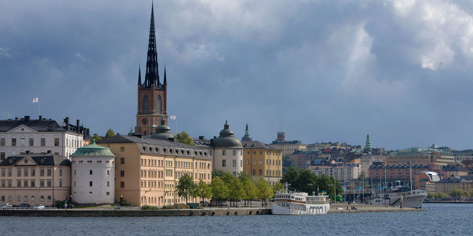 Holme med solbelysta hus och spetsigt kyrktorn bakom. I bakgrunden syns regntunga moln.