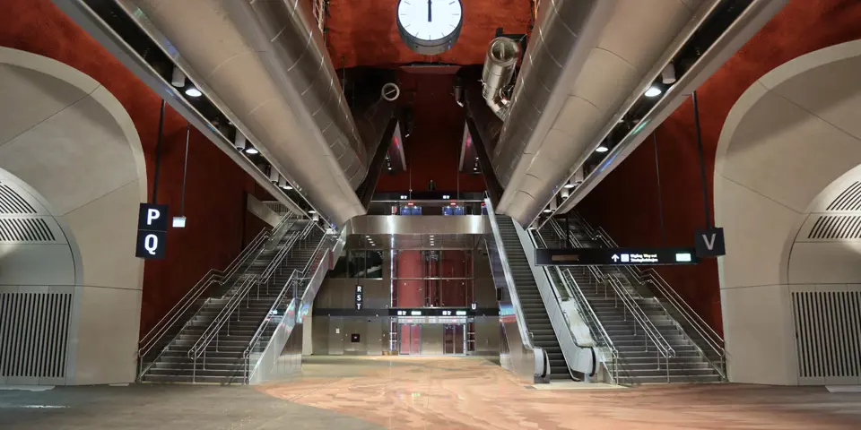 Vy upp mot trappor och rulltrappor i en vänthallsmiljö med rött golv och röda väggar. På båda sidorna framför trapporna finns valv med entréer.