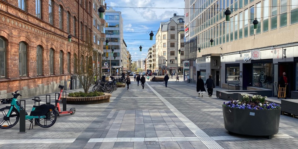 En gågata med byggnader på båda sidor. Cykelställ, stora krukor med blommor och människor i rörelse, foto.
