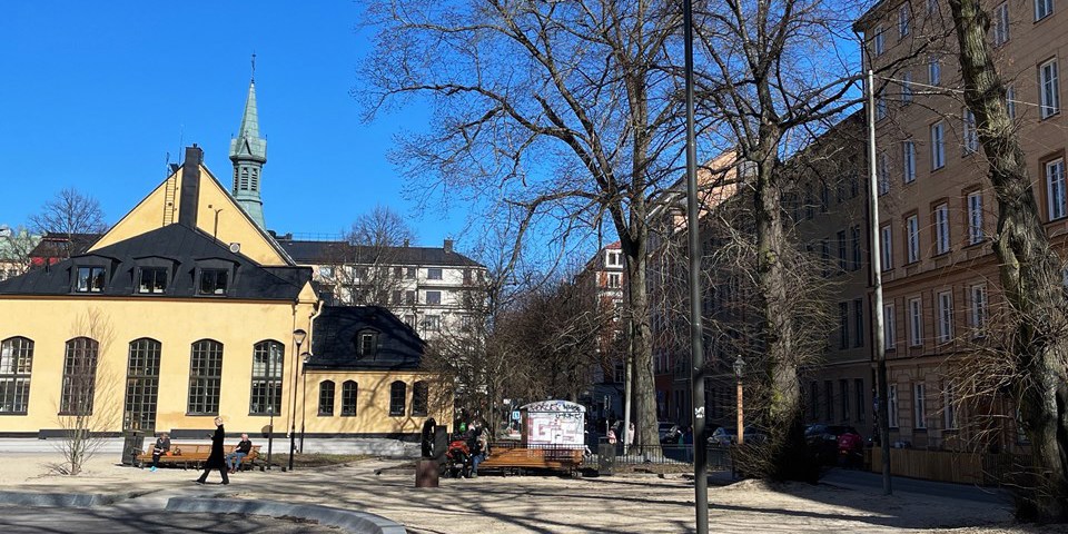 Parkyta omsluten av hus och träd. Några människor som sitter på bänkar och några som promenerar, foto.