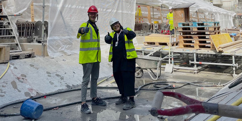 Två personer iklädda gula varselvästar och bygghjälmar står inne på en byggarbetsplats. De är glada och gör tummen upp mot kameran.
