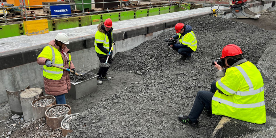 Två personer med gula västar och bygghjälmar står inne på byggarbetsplats, på varsin sida om en låda av plåt. Personerna håller i varsin spade och lägger grus ovanpå lådan. Framför de två personerna och lådan sitter två personer, också iklädda gula västar och bygghjälmar. De fotar händelsen.