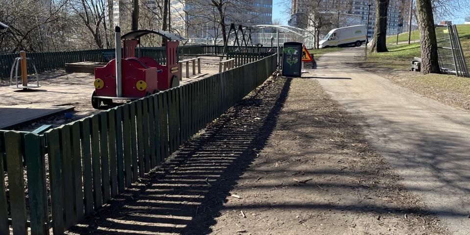 En gångväg går längs med en inhägnad lekplats med ett lektåg, sandlåda och gungor.
