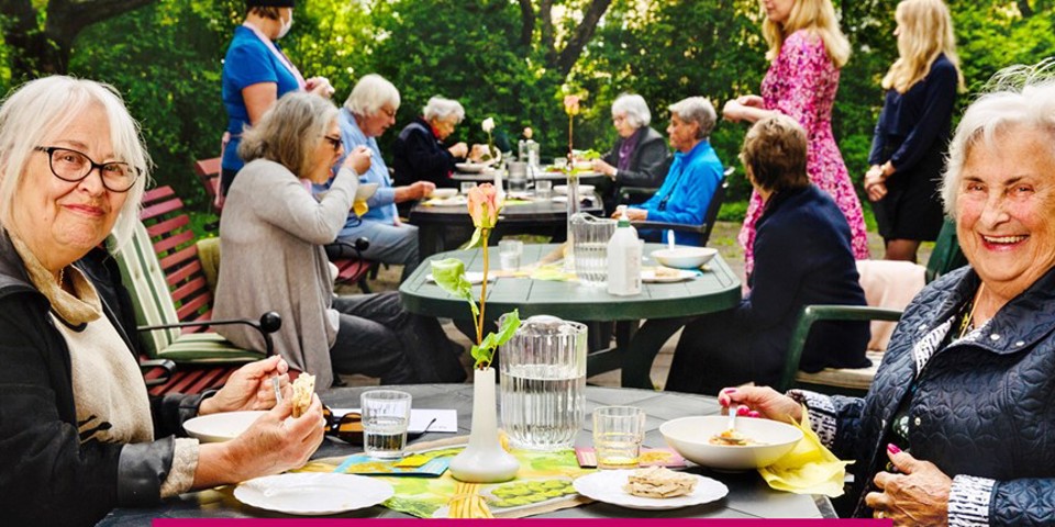 Två äldre personer sitter i en trädgård, andra människor syns i bakgrunden