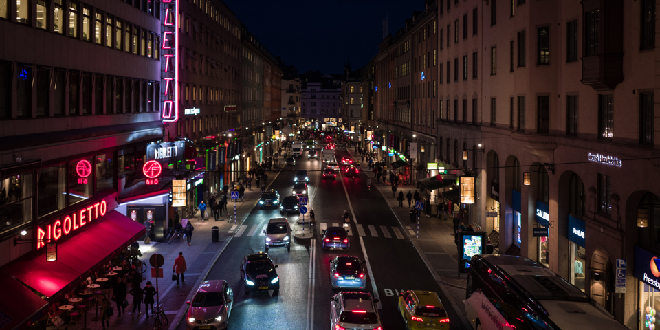 Kungsgatan kvällstid. Männiksor går och bilar kör. Neonskyltar lyser på husen.