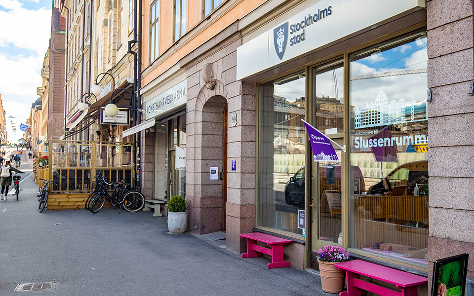Fasad utanför utställningslokalen Slussenrummet. Skyltfönster och entré med rosa bänkar och en blomkruka med rosa blommor utanför. Vid entrén hänger en lila flagga med texten "Öppet". Ovanför entrén sitter en skylt med Stockholms stads logo.