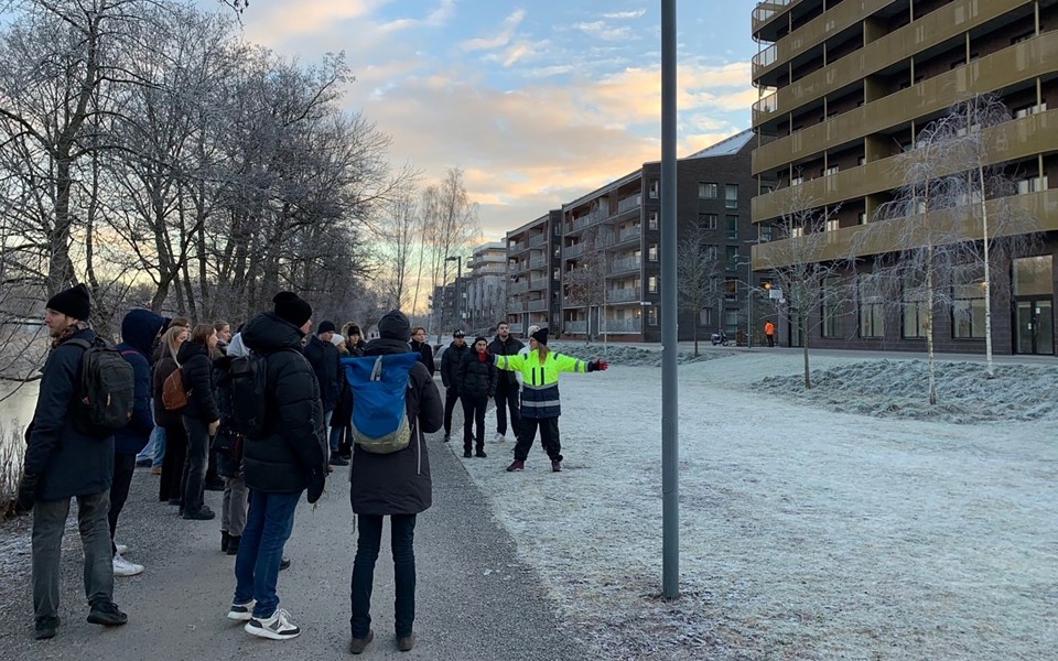 En grupp människor som står på en gräsmatta. Hus till höger i bild. 