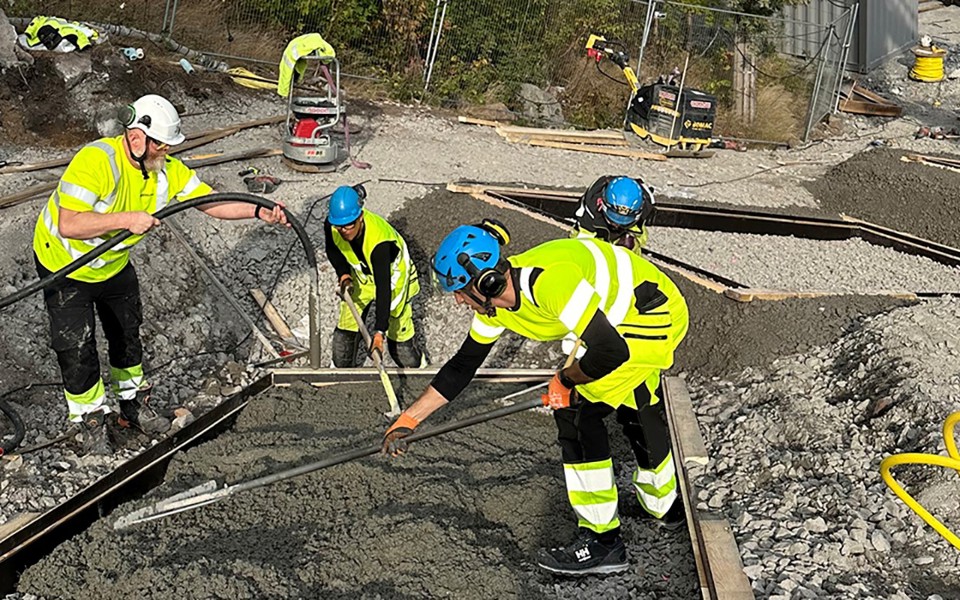 Tre byggarbetare i gula kläder häller cement i en stor form i en backe. 