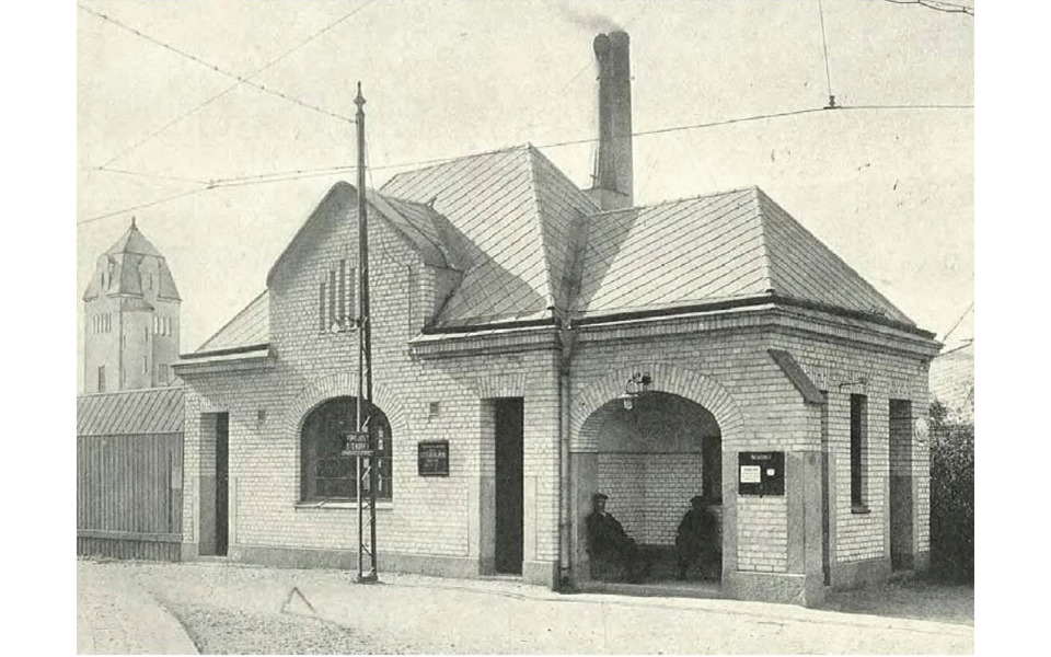 En historisk svartvit bild av en liten tegelbyggnad med valvbågar, tagen år 1914. Byggnaden har ett brant tak med en skorsten, och två personer sitter under en täckt ingång.