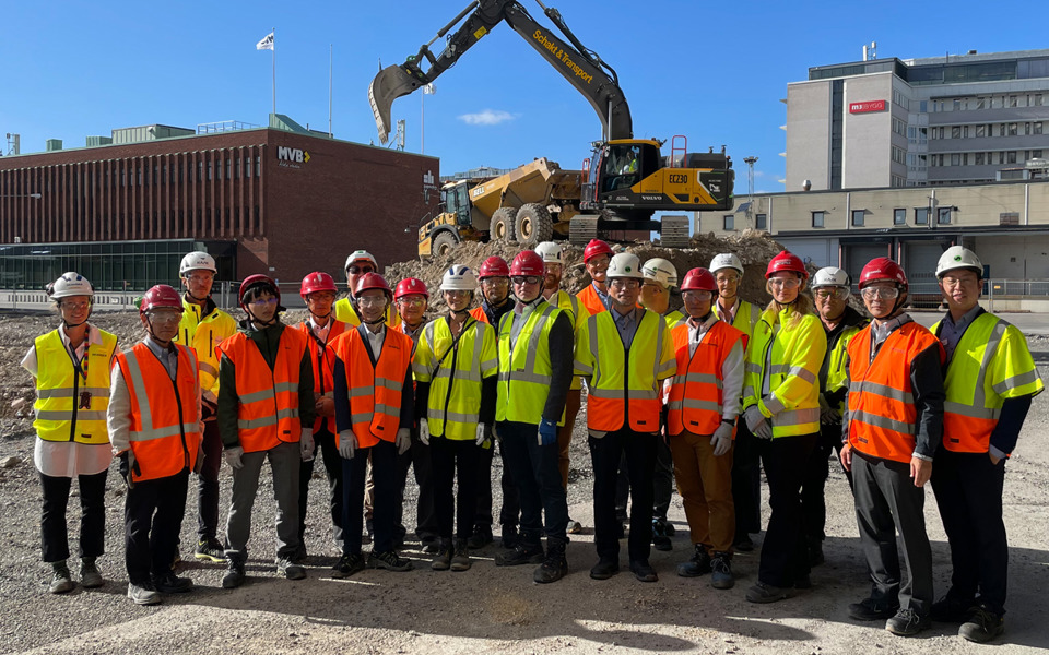 En grupp person på besök i Slakthusområdet med byggnader och en grävskopare i bakgrunden.