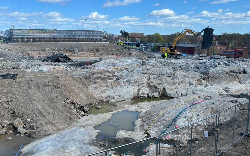En byggarbetsplats i Slakthusområdet där den andra och sista etappen av marksänkning pågår. Maskiner och utrustning används för att gräva och schakta bort massor. I bakgrunden syns byggnader och en stor arena under klarblå himmel med lite moln.