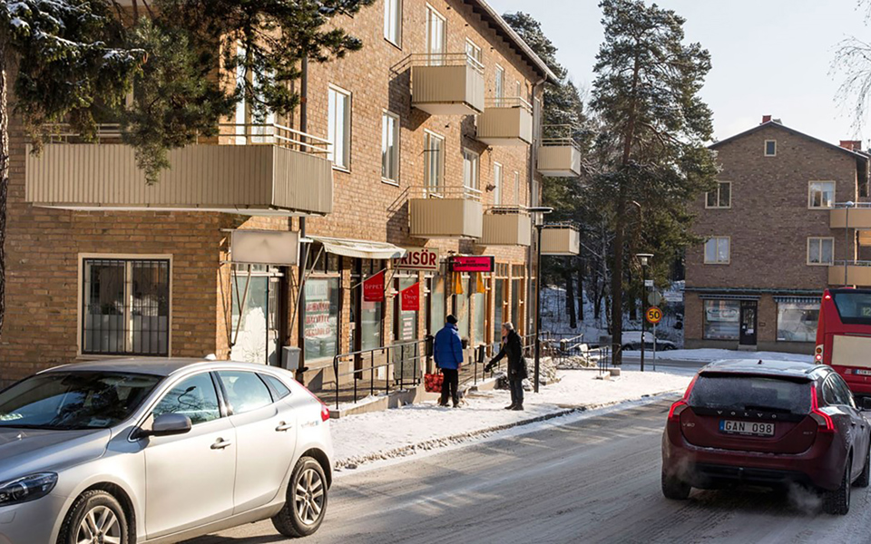 Bostadshus i tegel med frisörverksamhet på markplan. Parkerade bilar, omgivande träd och människor som samtalar. Foto.