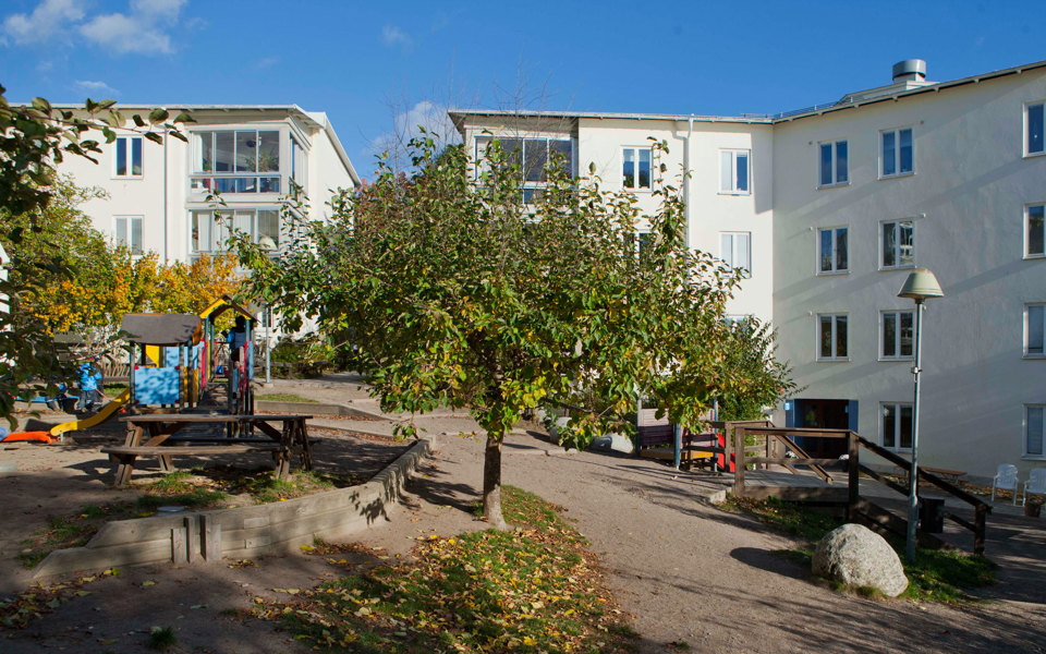 Innergård med lekplats, träd och bänk med bord. Omgiven av fler bostadshus, foto.
