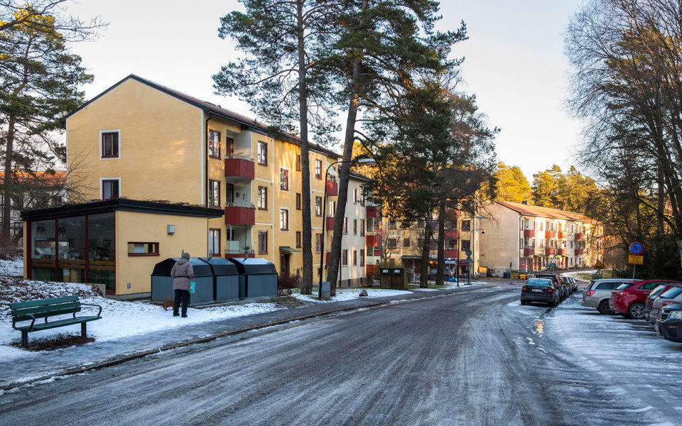 En gata i bostadsområde kantad av flerbostadshus och träd.  Bilar finns parkerade på gatan och parkeringsplats, foto.