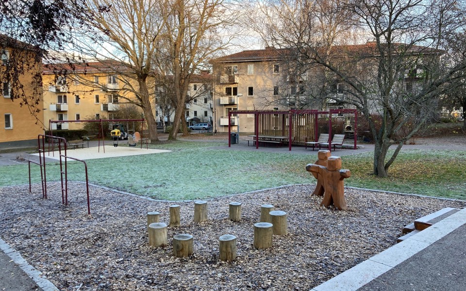 Lekplatsen Bomullen ligger i ett bostadsområde med gula och beigea flerfamiljshus. På lekplatsen finns en gräsmatta, flera träd och olika lekytor med gungor, små trädstubbar för de små barnen att sitta på och andra sittmöjligheter.