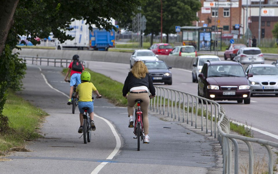 Cyklande barn och vuxna på cykelbana bredvid bilväg. Bilar i rörelse, foto.