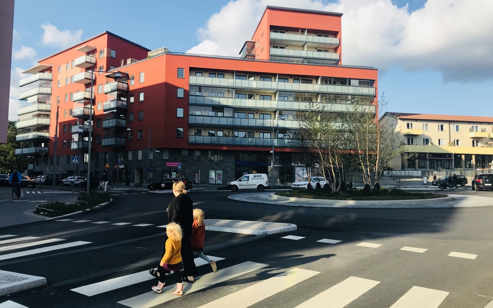 Bostadshus i olika nivåer med verksamhetslokaler på bottenplan, bakom en rondell. Människor i rörelse, fotografi.