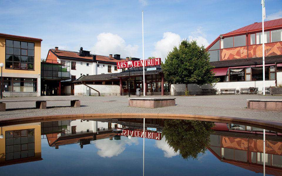 Ett stenbelagt torg med med en damm och sittplatser. Omslutet av hus, bibliotek och kafé, foto. 