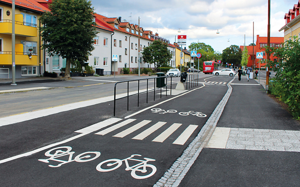 En bilväg med cykelbana. Vägen kantas av flerbostadshus, träd och buskar, foto.