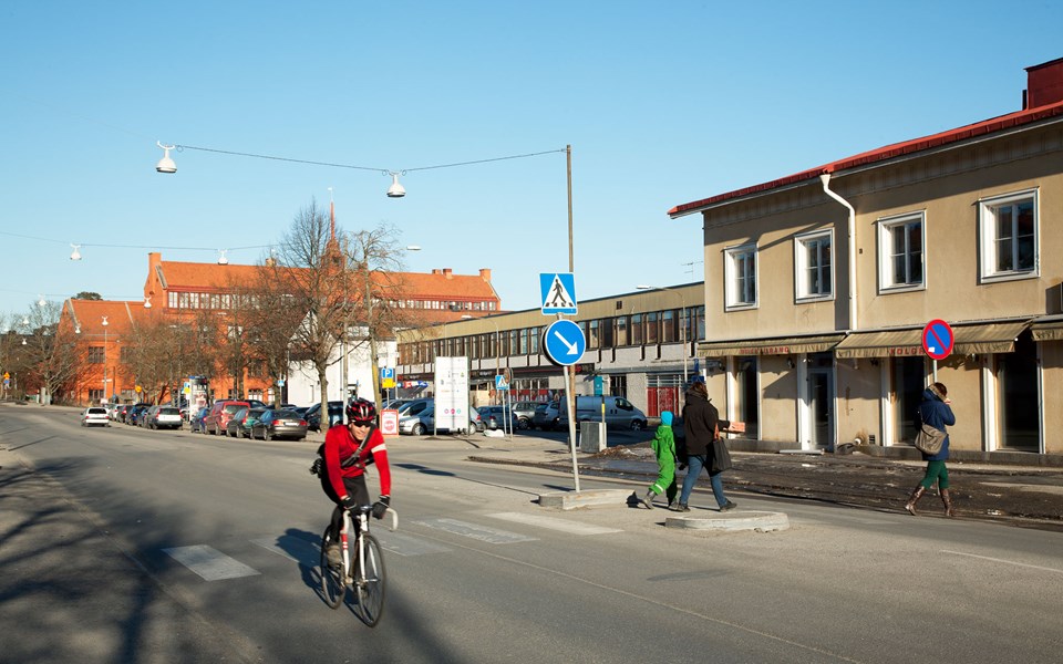 En bilväg genom bostadsområde med övergångsställe. En cyklist och människor som korsar vägen, foto.