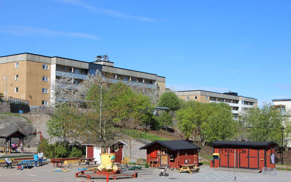 Solig dag i parklek med röda småhus. Träd och brungrå flervåningshus i bakgrunden, foto.