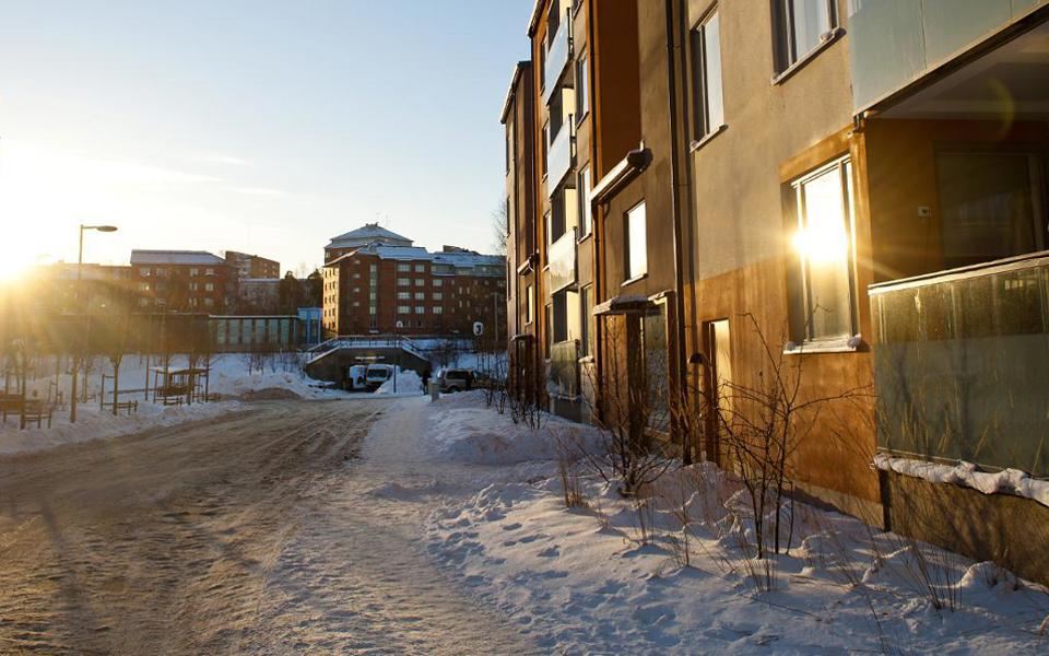 En gata med flerbostadshus på vardera sidan och med en pendeltågsstation i bakgrunden. Det är vinter och gatan är snöröjd. Solen ligger lågt och skiner mellan husen. Foto.