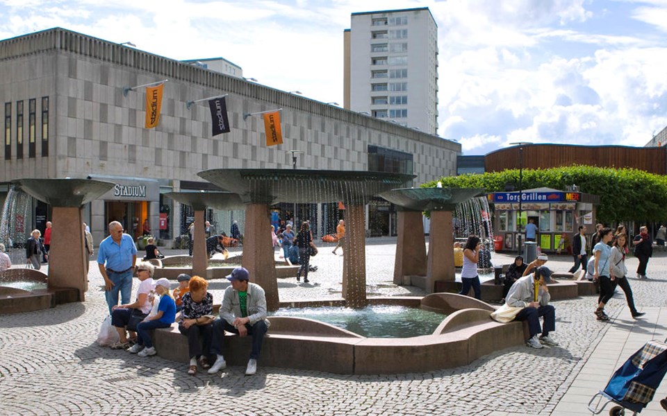 En torgyta med fyra fontäner omgiven av butiker. På torget finns en grillkiosk  och människor i rörelse. Foto.