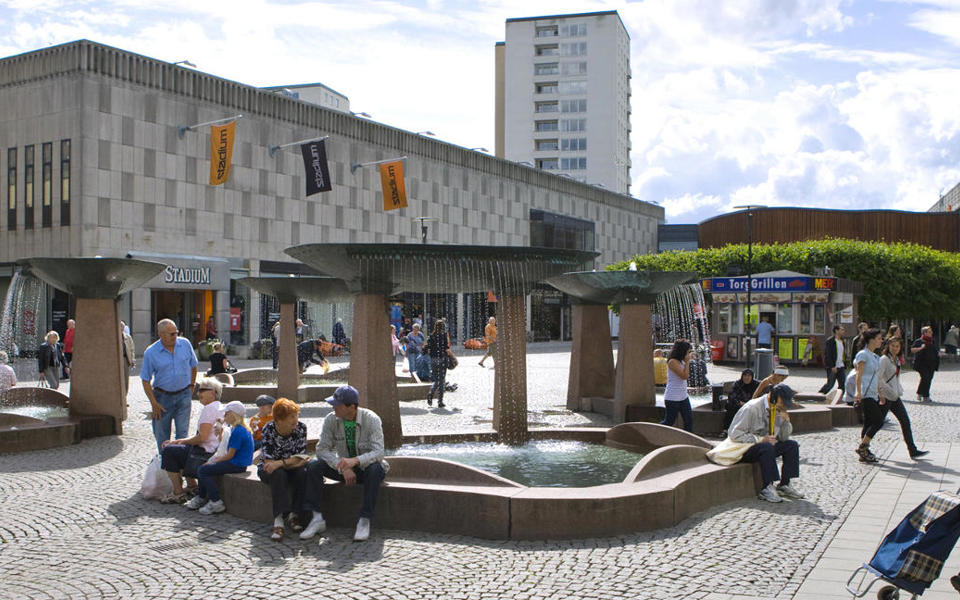 En torgyta med fyra fontäner omgiven av butiker. På torget finns en grillkiosk  och människor i rörelse. Foto.